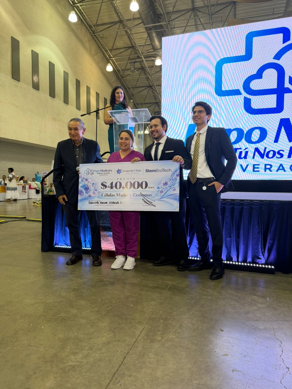 La ganadora con el cheque del premio, acompañada por el equipo de Longevity Clinic, frente a la pantalla de la Expo Médica Tú Nos Inspiras